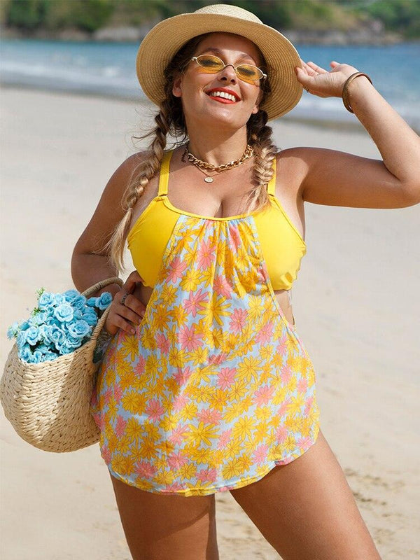Une femme ronde pose sur la plage en tankini. Le haut est un bandeau jaune cousu à un top ajouré sur les côtés au motif liberty bleu, rose et jaune. Le top est évasé. Le bas est une culotte jaune. 