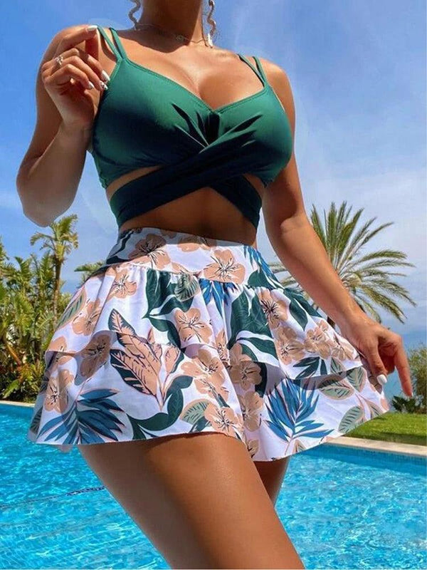 Une femme pose au bord d'une piscine avec un tankini. Le haut est un top croisé vert. Le bas est une jupe blanche à motif tropical. La jupe est taille haute, cintrée à la taille et évasé ensuite pour donner un effet volant.